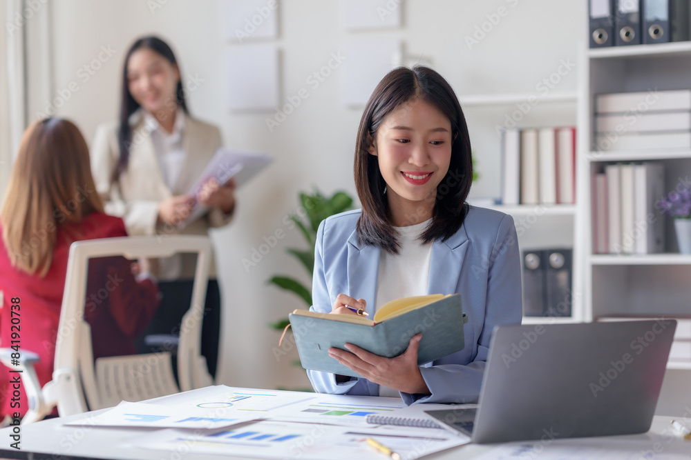 Smiling businesswoman working on laptop in a modern office, with colleagues in the background, discussing projects and documents.