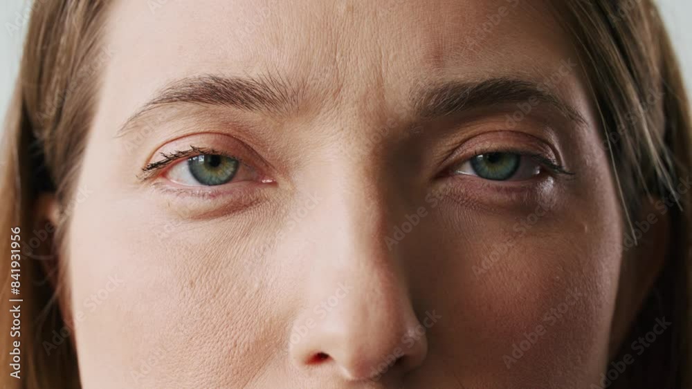 Very close camera view of Caucasian girls face. Woman with charming green eyes and straight brown hair. Not expressing any emotions. Wearing makeup. Looking directly at camera. Blinking eyes.