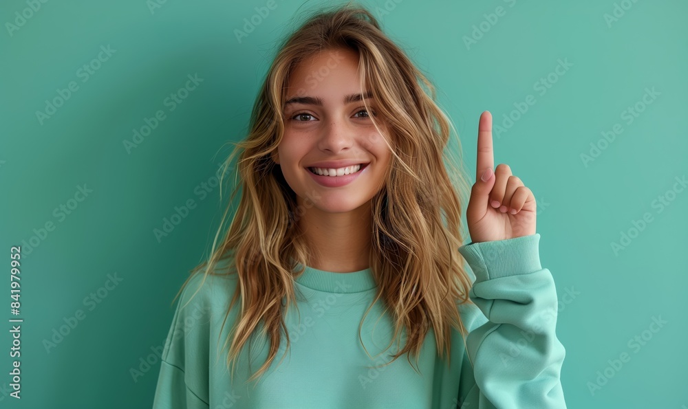 Smiling caucasian teenager girl in a mint sweatshirt smiling with healthy teeth and pointing index finger up at copy space expressing wow emotion standing isolated on paper mint background Smiling caucasian teenager girl in a mint sweatshirt smiling with healthy teeth and pointing index finger up at copy space expressing wow emotion standing isolated on paper mint background