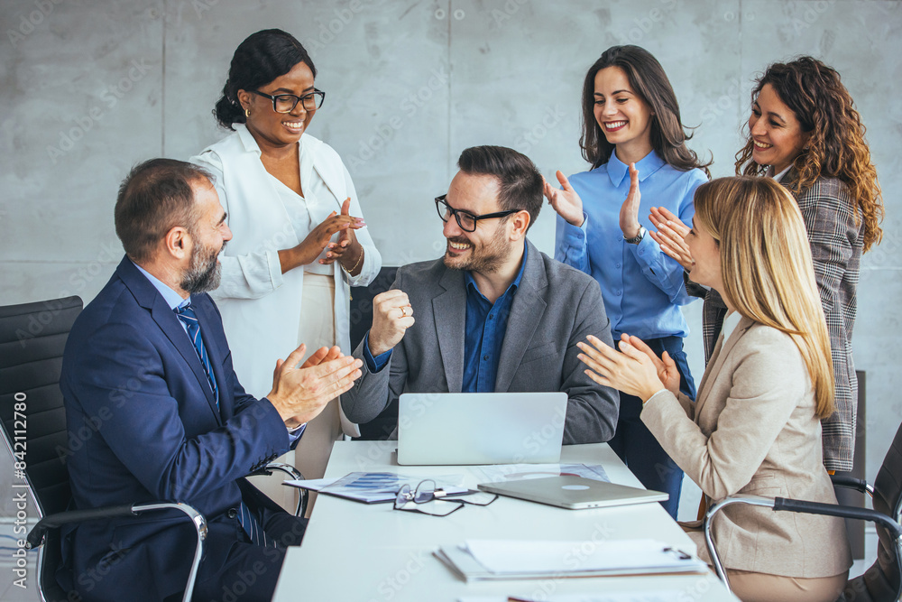 © Dragana Gordic - A group of diverse business professionals celebrate success, clapping for a smiling male colleague seated with a laptop in a modern office.