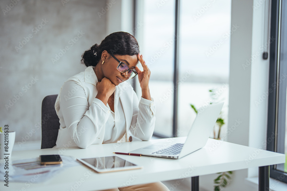 A professional African American businesswoman concentrates on her laptop in a modern office, her attire and glasses reflecting corporate style.