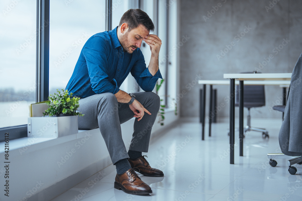 A weary Caucasian businessman sits in a corner, rubbing his forehead, reflecting the stress of his profession surrounded by a sleek office environment. A weary Caucasian businessman sits in a corner, rubbing his forehead, reflecting the stress of his profession surrounded by a sleek office environment.