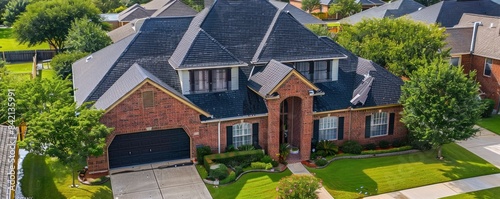 An aerial view of a residential roof with significant damage. Tiles are missing and broken, revealing underlying structures. The roof shows signs of wear and tear, set against a backdrop of lush trees