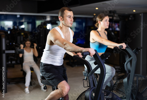 Determined young man and woman training with air-bike in fitness hall during full-body workout