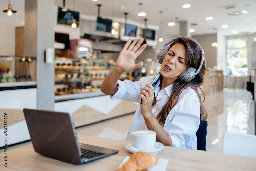 Beautiful and happy young woman sitting and eating delicious rolls in bakery or fast food. She also using her laptop and listening to music with headphones.