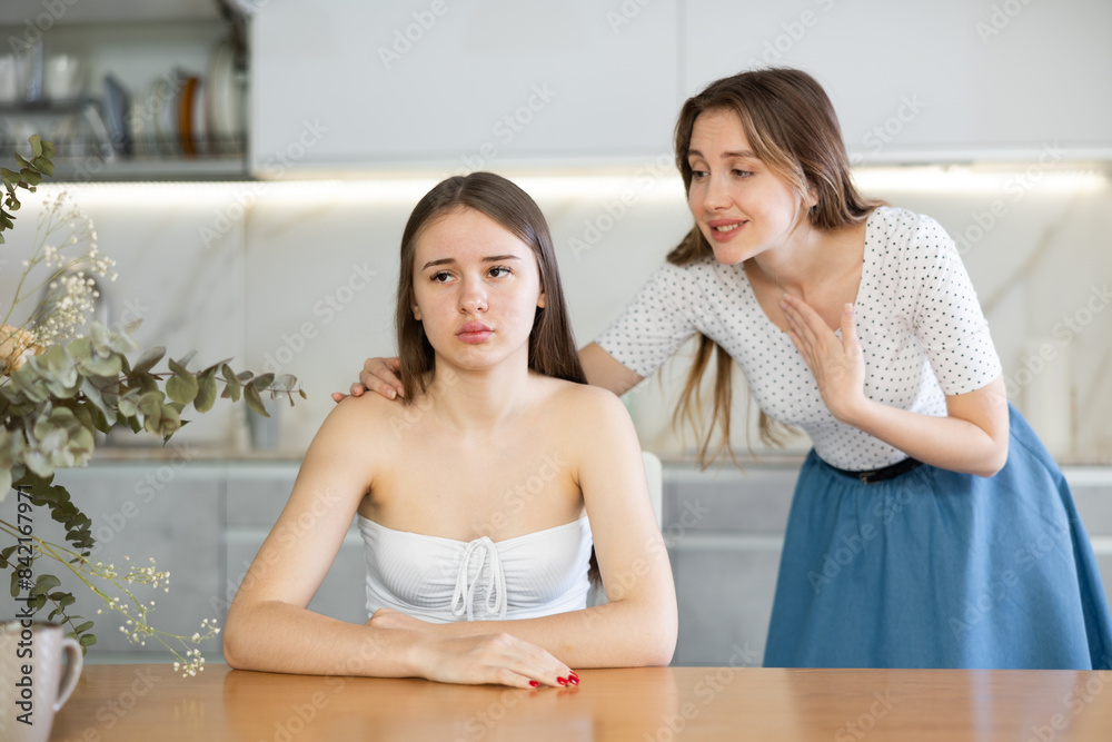 Obraz premium Upset teen girl sitting at the table, her mother speaking to her standing behind in the kitchen