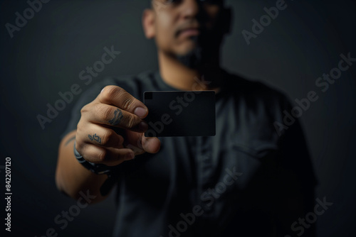 A close up image of a hispanic latinx man holding a black business card up to the camera, shallow depth of field. Isolated, black background. Black tee shirt.