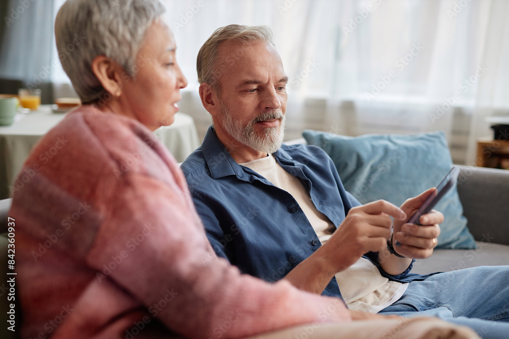 Side view portrait of modern senior couple using phone with mobile app together while sitting on couch at home Side view portrait of modern senior couple using phone with mobile app together while sitting on couch at home