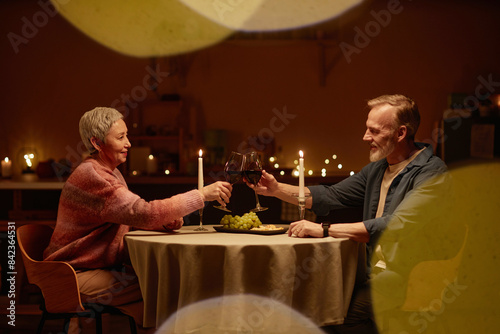 Side view portrait of senior couple enjoying romantic dinner at home and clinking wine glasses at candlelit table copy space photo