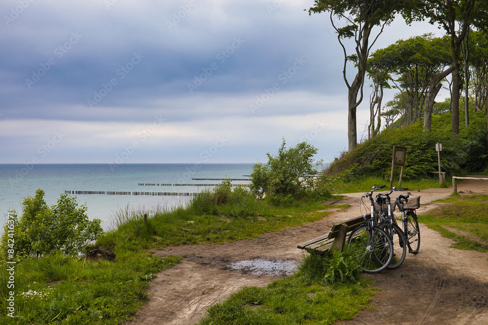 Fototapeta premium Fahrrad-Pause am Ostseeküsten-Radweg mit Blick vom Steilufer auf die Ostsee am Gespensterwald Nienhagen, Fahrräder parken an einer Bank am Kliff vor markanten Buchen unter Wolkenhimmel, Deutschland