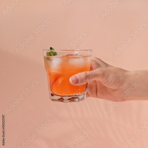 Refreshing peach fruit mocktail with ice and mint, hand holding glass with orange juicy soft drink on pastel pink background