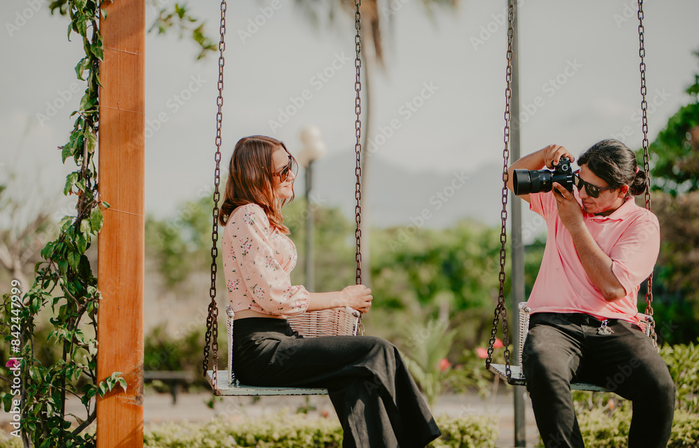 Obraz premium Boyfriend taking a photo of his girlfriend on a swing in a park. Boyfriend photographing girlfriend sitting on a swing