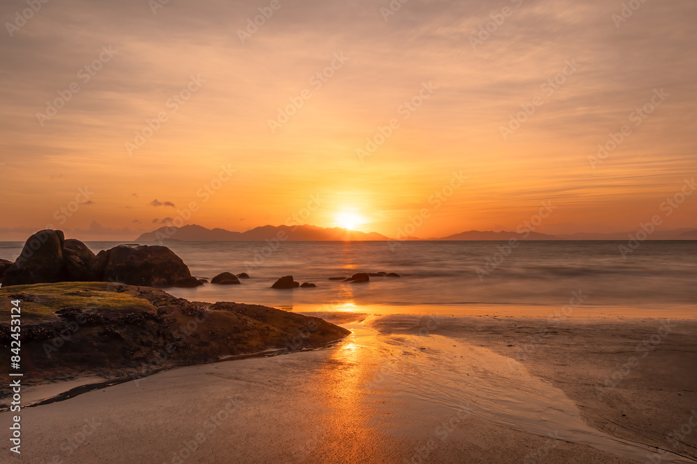 A glowing orange sunrise in a cloudy sky over the sea and beach with reflections in the wet beach sand next to dark, silhouetted granite rocks at Rose Bay in Bowen in tropical Queensland, Australia. A glowing orange sunrise in a cloudy sky over the sea and beach with reflections in the wet beach sand next to dark, silhouetted granite rocks at Rose Bay in Bowen in tropical Queensland, Australia.