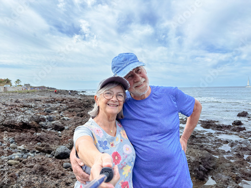 Happy smiling senior couple in retirement embracing at the beach enjoying sunny day at sea while taking self portraits together with a smartphone and selfie stick photo