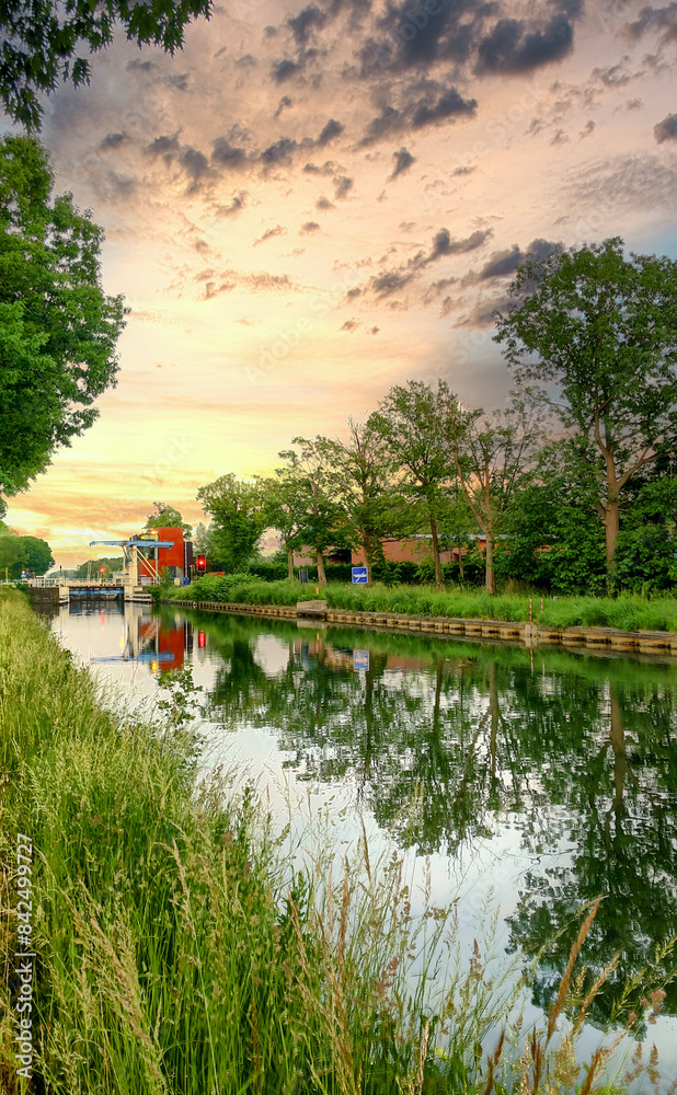 Fototapeta premium Idyllic sunset with beautiful reflections on a tranquil canal, surrounded by greenery and a cloudy sky.