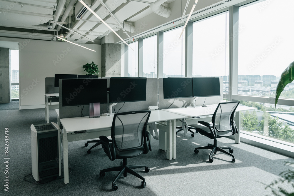 A modern, open-plan office space filled with natural light from large windows that offer a view of the cityscape. The office features a clean, minimalistic design with chairs and computer monitors. A modern, open-plan office space filled with natural light from large windows that offer a view of the cityscape. The office features a clean, minimalistic design with chairs and computer monitors.