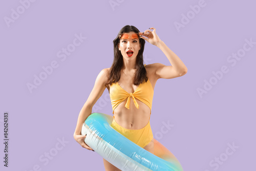 Surprised young woman in beachwear with swim ring on lilac background