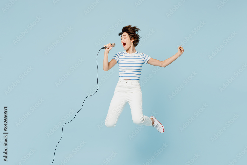 Full body young surprised shocked woman wears striped t-shirt casual clothes jump high sing song in microphone isolated on plain pastel light blue cyan background studio portrait. Lifestyle concept. Full body young surprised shocked woman wears striped t-shirt casual clothes jump high sing song in microphone isolated on plain pastel light blue cyan background studio portrait. Lifestyle concept.