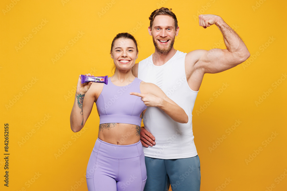 Young strong fitness trainer instructor sporty two man woman wearing blue clothes spend time in home gym hold in hand protein energy bar isolated on plain yellow background. Workout sport fit concept. Young strong fitness trainer instructor sporty two man woman wearing blue clothes spend time in home gym hold in hand protein energy bar isolated on plain yellow background. Workout sport fit concept.