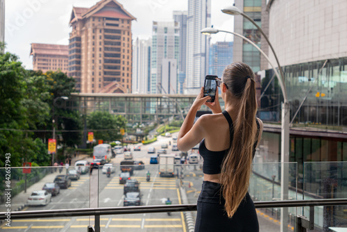 Photography A woman in a black top photographs the Kuala Lumpur skyline from a glass bridge, capturing high-rise buildings and bustling city life
