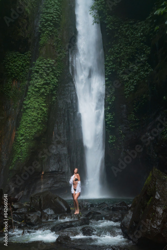 A woman in a black bikini and white shirt stands on rocks near a tall waterfall  surrounded by lush greenery.
