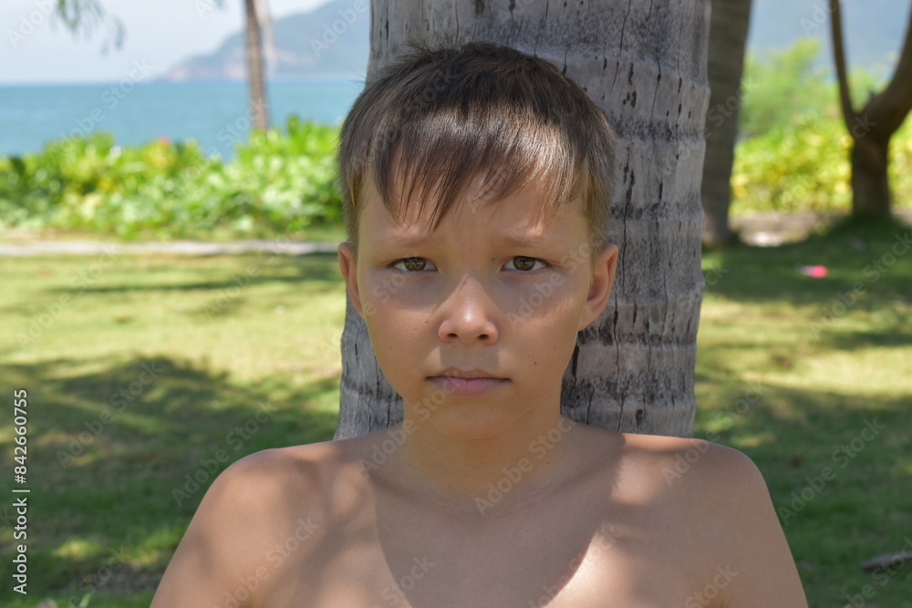 A blond boy sits in the shade of a palm tree and looks at the camera.