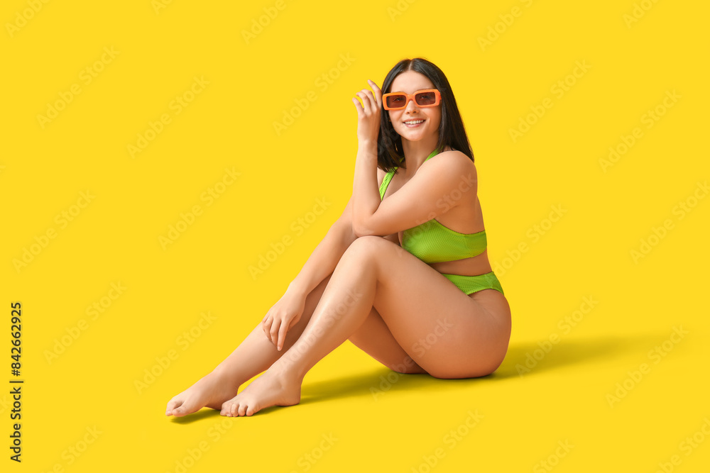 Beautiful young woman in stylish green swimsuit and sunglasses sitting on yellow background Beautiful young woman in stylish green swimsuit and sunglasses sitting on yellow background