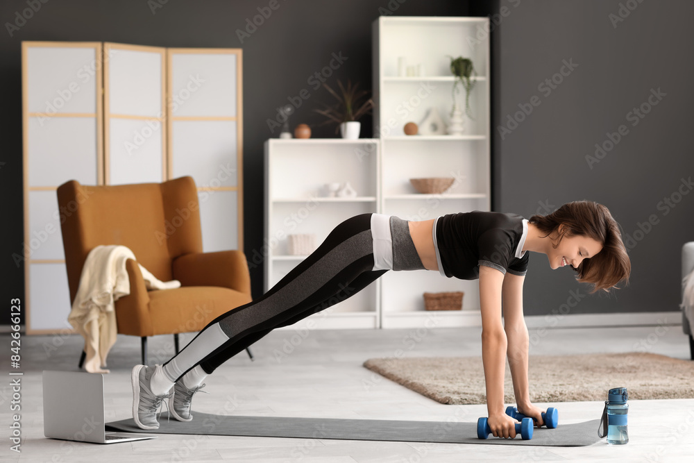 Beautiful young woman training with dumbbells and laptop on yoga mat in living room