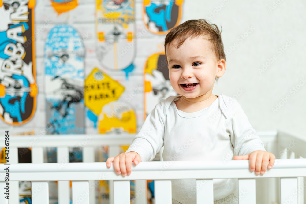 A smiling baby in a white shirt with the word ninja on it. The baby is standing in front of a crib with a skateboard on the wall A smiling baby in a white shirt with the word ninja on it. The baby is standing in front of a crib with a skateboard on the wall