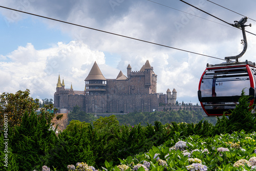 A scenic view of a medieval-style castle at Ba Na Hills in Danang, Vietnam. The castle features tall towers and a cable car passing by, with green foliage and cloudy sky in the background.