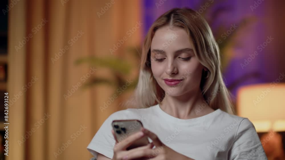 A young woman is smiling while using a smartphone in a warm and cozy home setting A young woman is smiling while using a smartphone in a warm and cozy home setting