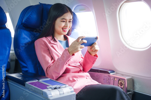 A young Asian female airplane passenger sits by the window during the flight, holding her boarding pass, with her carry-on luggage stored overhead, wearing her seatbelt, and enjoying the journey.