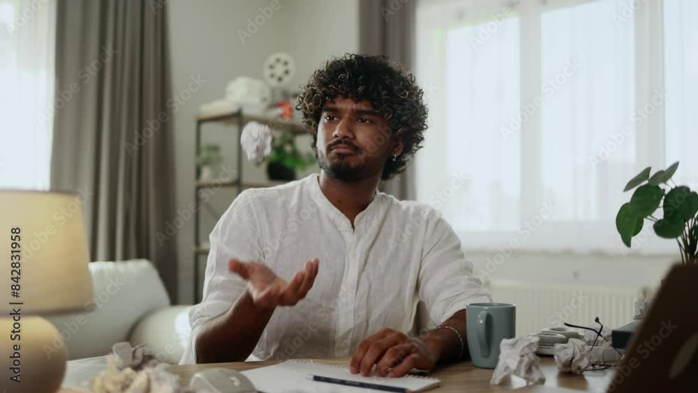 Tired young handsome indian man student or freelancer thinks questioningly throwing up crumpled paper while finding solution and writing on paper at home workplace Tired young handsome indian man student or freelancer thinks questioningly throwing up crumpled paper while finding solution and writing on paper at home workplace