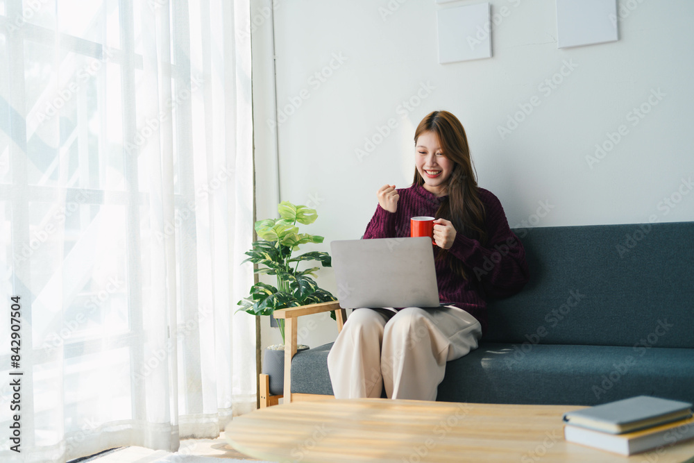 Asian woman is sitting on a couch with a laptop and a cup of coffee Asian woman is sitting on a couch with a laptop and a cup of coffee