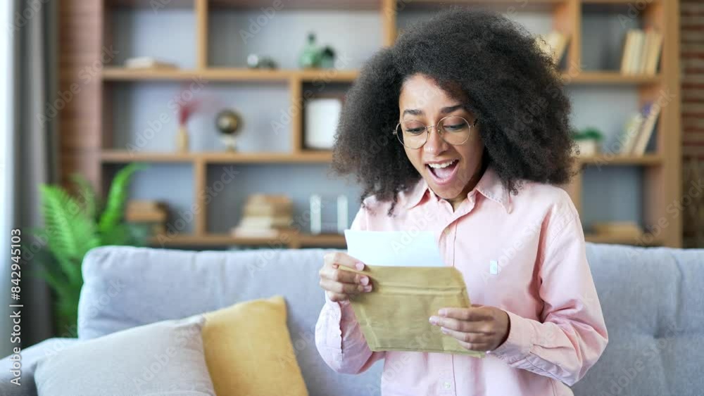 Happy excited female university student celebrating success reading letter with good news while sitting on sofa in room at home. Smiling lady applicant holds joyful notice of successful entrance exams