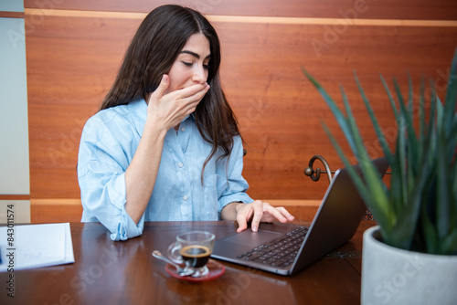 tired young businesswoman covering her mouth while working on laptop at desk