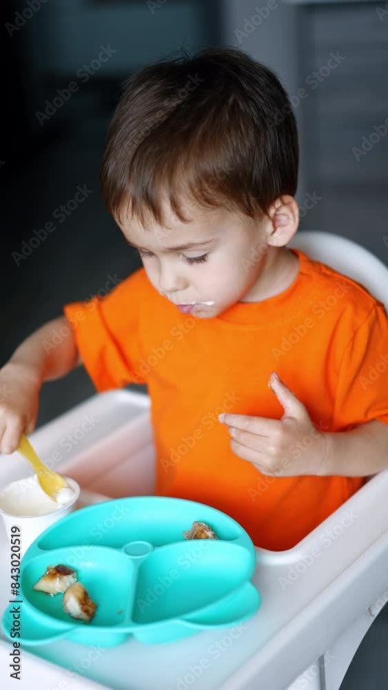 Adorable Caucasian baby using spoon to eat some yoghurt from a cup. Kid drops some food and looks at it on the floor. Vertical video. Adorable Caucasian baby using spoon to eat some yoghurt from a cup. Kid drops some food and looks at it on the floor. Vertical video.
