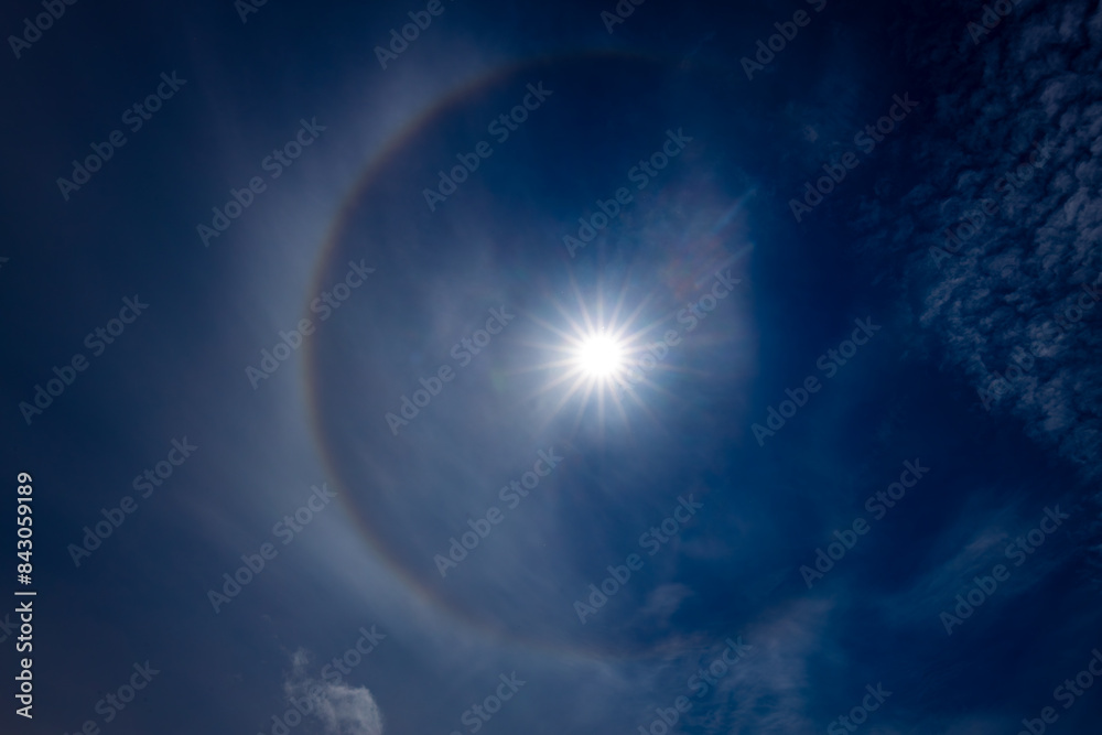 A captivating image of a sun halo against a deep blue sky, created by the refraction of sunlight through ice crystals in the atmosphere. The sun is bright, surrounded by a circular rainbow-like halo. A captivating image of a sun halo against a deep blue sky, created by the refraction of sunlight through ice crystals in the atmosphere. The sun is bright, surrounded by a circular rainbow-like halo.