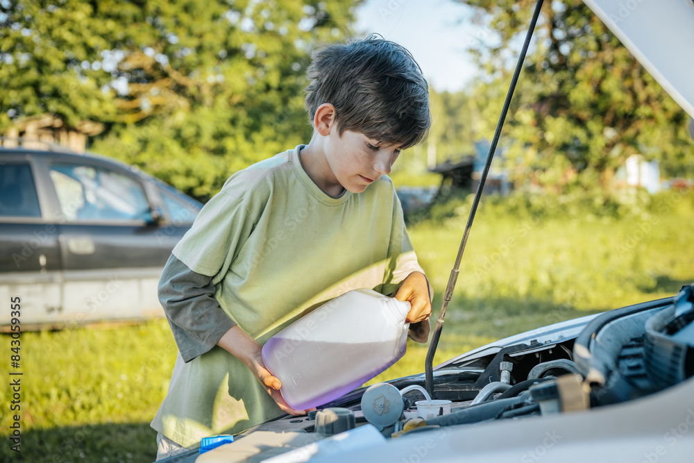 Fototapeta premium A young boy pours windshield washer fluid into a car's engine compartment on a sunny day. The scene is set outdoors with a blurred green background.