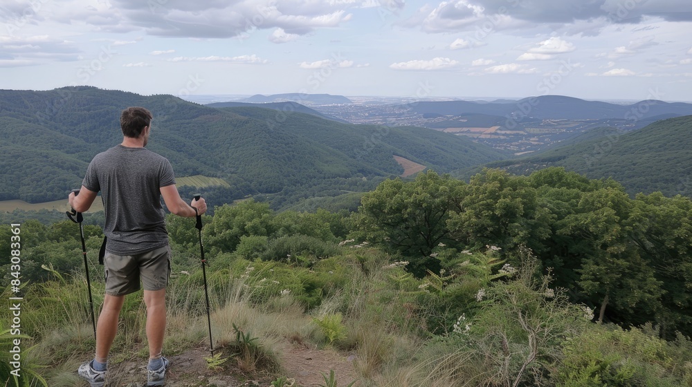 A man with hiking sticks stands at the top of a mountain and looks at a forest landscape, mountains and sky in the distance. A man with hiking sticks stands at the top of a mountain and looks at a forest landscape, mountains and sky in the distance.