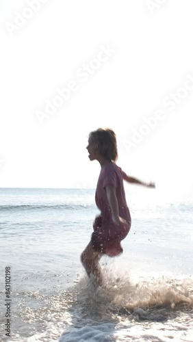 Vertical video. Little girl pink dress having fun while jumping in sea water, slow motion. Happy childhood and family vacation concept 