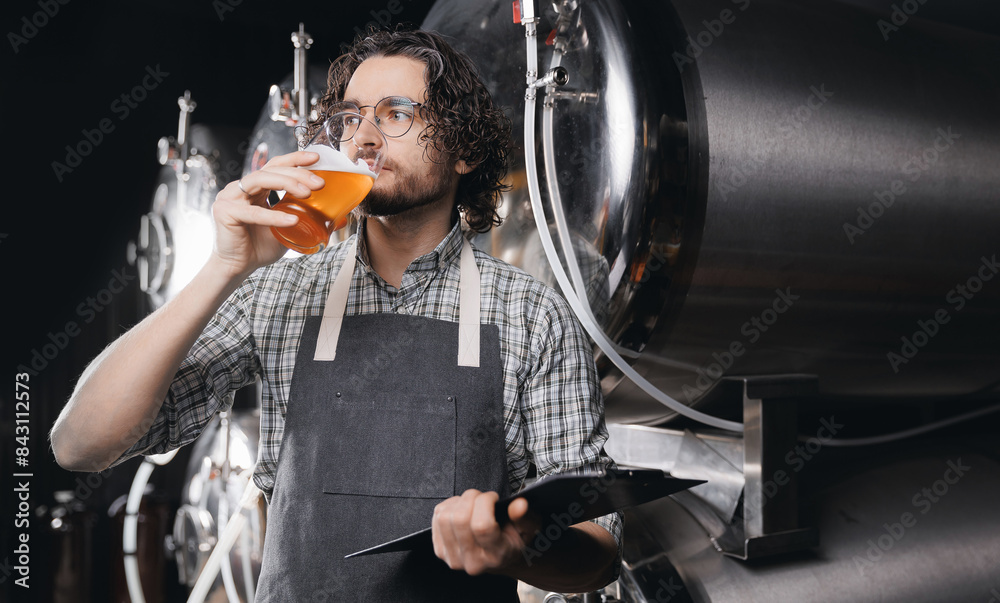 Fototapeta premium Expert brewer with clipboard in apron holds glass of craft beer and checking quality and color. Worker sommeliers taste drink on Brewery factory