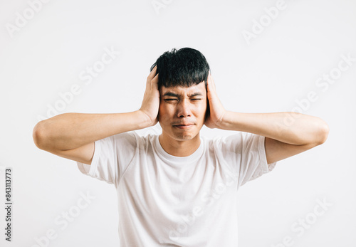 An Asian man in pain, using his hands to cover his ears and shutting his eyes to cope with the loud noise. Studio shot isolated on white background, showing his need for relief.