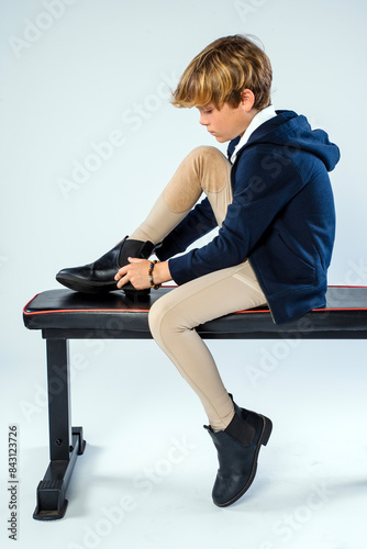 Young male horse rider in equestrian clothing sitting on a bench putting on one of his riding boots photo