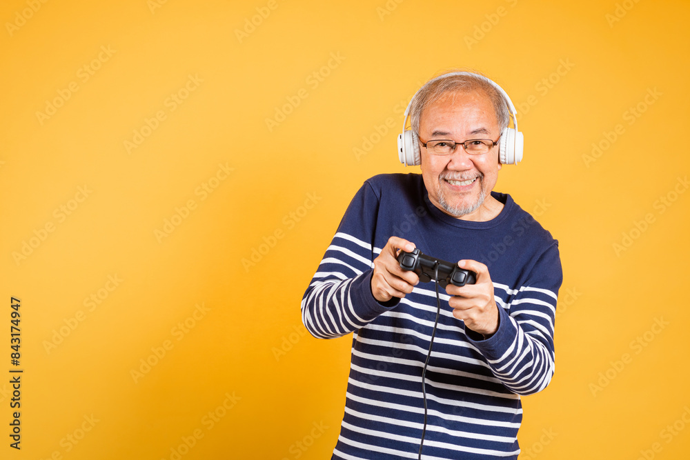Portrait Asian smiling old man with headphones holding game controller studio shot isolated yellow background, senior man pensioner feeling winning playing a video game with joystick console Portrait Asian smiling old man with headphones holding game controller studio shot isolated yellow background, senior man pensioner feeling winning playing a video game with joystick console