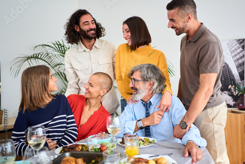 Joyful Caucasian family smiling looking at each other hugging and having fun indoor. People of diverse ages gathered at table of food for enjoy meal together at home