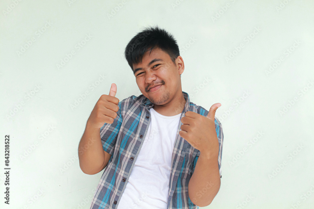 Excited and happy Asian man gives thumbs up hand gesture of approval, isolated by gray background.