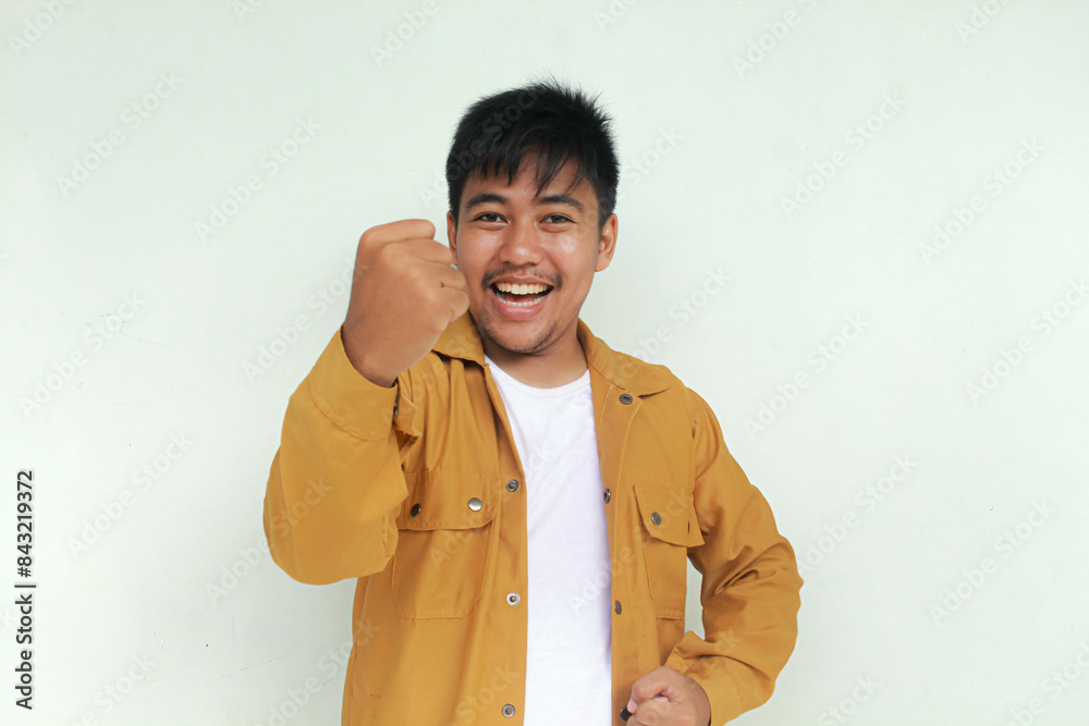 Happy excited and smiling young Asian man raising his arm up and and scream to celebrate success or achievement. Isolated in gray background.