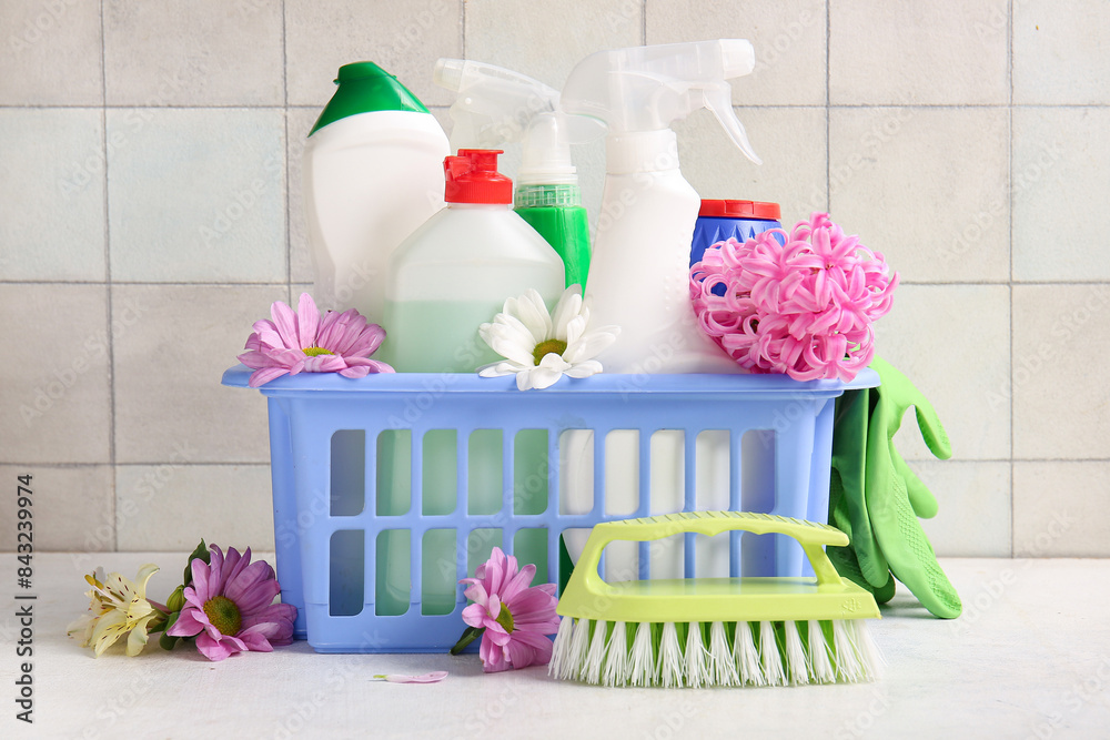 Basket with set of cleaning supplies and spring flowers on white table against tiled background Basket with set of cleaning supplies and spring flowers on white table against tiled background