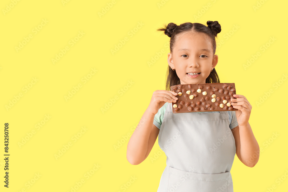 Cute little Asian girl in apron with chocolate bar on yellow background Cute little Asian girl in apron with chocolate bar on yellow background